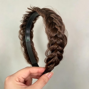 Puede incluir: Una diadema trenzada de color marrón con cabello sintético tejido en una trenza gruesa y texturizada. El cabello es de un tono marrón medio con reflejos sutiles, unido a un marco de diadema de plástico negro con dientes internos.