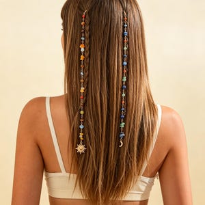 Könnte beinhalten: Zwei lange Haarketten mit bunten Perlen, kleinen Pilzanhängern sowie goldfarbenen Metallanhängern in Form einer Sonne und einer Mondsichel. Die Ketten sind an braunen Haarsträhnen befestigt, wobei eine Kette in einen Zopf eingearbeitet ist.