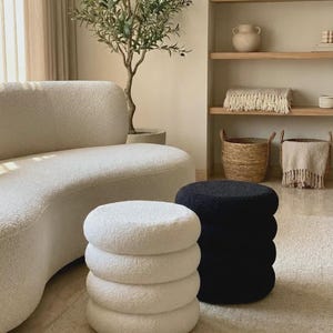 May include: Two cylindrical ottomans with a textured, nubby fabric finish. One ottoman is solid white and the other is solid black. Both feature a stacked, rounded ring design that creates a tiered appearance. These pieces are floor-standing.