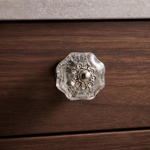 May include: A clear, octagonal glass cabinet knob with a crackled texture and a central silver-toned metal floral accent, mounted on a dark brown wooden drawer front with a visible horizontal wood grain pattern and a smooth finish.