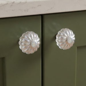 May include: Two round, clear glass cabinet knobs with a fluted, scalloped edge design. Each knob features a central circular indentation and is mounted onto a flat-panel cabinet door painted in a solid, matte olive green color.
