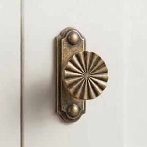May include: A circular, fluted brass cabinet knob with a sunburst pattern, mounted on a rectangular brass backplate with rounded ends and two visible screw heads. The hardware is attached to a smooth, matte white cabinet door surface.