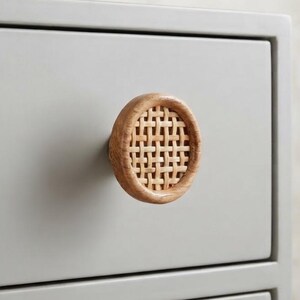 May include: A circular drawer knob featuring a light brown woven rattan center encased in a smooth wooden frame, mounted on a flat, matte light gray drawer front. The knob has a textured, grid-like pattern and a natural wood grain finish.