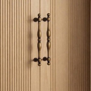 May include: A light-colored wooden cabinet door featuring a vertical fluted or ribbed texture. Two matching antique brass-toned metal handles are mounted vertically side-by-side in the center, each with a slender, rounded, and contoured shape.