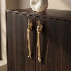 May include: A dark brown wooden cabinet featuring two gold-toned metal cabinet pulls shaped like figures sitting with their legs dangling down. A textured, off-white, organic-shaped ceramic vase sits on the top surface of the cabinet.
