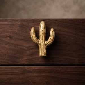 May include: A gold-toned metal cabinet knob shaped like a saguaro cactus with a textured surface. The cactus knob is mounted onto a dark brown wooden drawer front with a visible horizontal grain pattern and a clean horizontal seam line.