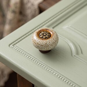 May include: A round ceramic cabinet knob with a speckled cream and brown glaze finish. The center features a circular antique brass-toned metal inlay embossed with a floral petal design. The knob is mounted on a light sage green paneled surface.