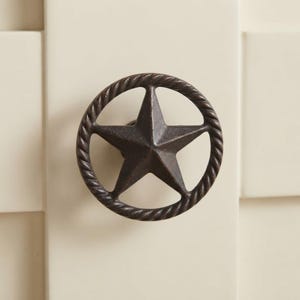May include: A dark brown metal cabinet knob featuring a five-pointed star centered within a circular frame. The outer ring of the circle has a twisted rope texture, and the entire piece has a matte, slightly weathered finish with dark tones.