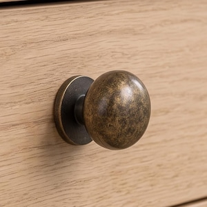 May include: A round cabinet knob with a dark, distressed antique brass finish and a matching circular backplate, mounted onto a light-colored wooden drawer front with a visible horizontal grain pattern throughout the surface.