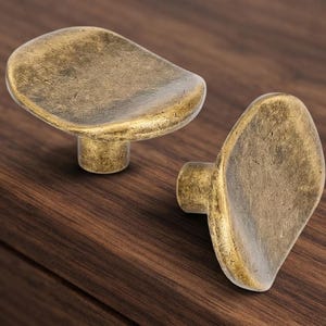 May include: Two cabinet knobs with an irregular, organic shape and a textured, antique brass finish. Each knob features a wide, slightly concave top surface and a cylindrical base, showcasing a weathered metallic appearance with dark highlights.