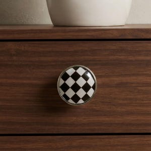 May include: A round cabinet knob featuring a black and white checkered pattern. The knob has a smooth, glossy finish and is framed by a thin, polished silver-toned metal border, mounted onto a dark brown wood-grained drawer front surface.