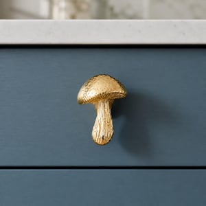 May include: A gold-colored mushroom-shaped cabinet knob with a textured, metallic finish. The knob features a rounded cap and a vertical stem, mounted onto a flat, dark blue drawer front surface with a horizontal seam visible below the knob.