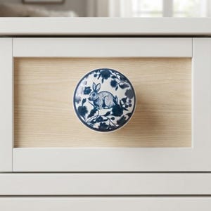 May include: A round ceramic cabinet knob featuring a white background with a dark blue floral pattern and a central illustration of a rabbit. The knob is mounted on a light-colored wood panel framed by a white border on a piece of furniture.
