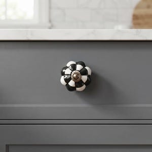 May include: A decorative cabinet knob featuring a scalloped, flower-like shape with a high-gloss finish. The surface displays a repeating black and white checkered pattern, centered with a small, circular metallic bronze-toned hardware piece.