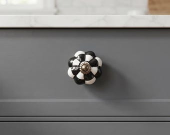 May include: A decorative cabinet knob featuring a scalloped, flower-like shape with a high-gloss finish. The surface displays a repeating black and white checkered pattern, centered with a small, circular metallic bronze-toned hardware piece.