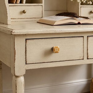 May include: A square, mustard-yellow ceramic drawer knob with a white floral pattern, mounted on a distressed cream-colored wooden desk. The knob features a central metal screw attachment and a smooth, glossy finish on its front surface.