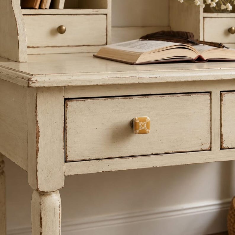 May include: A square, mustard-yellow ceramic drawer knob with a white floral pattern, mounted on a distressed cream-colored wooden desk. The knob features a central metal screw attachment and a smooth, glossy finish on its front surface.
