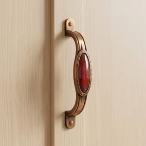 May include: A curved brass-toned metal cabinet handle featuring a central oval inlay of polished red stone. The handle is mounted with two screws and displays a decorative Greek key pattern border surrounding the smooth red center piece.