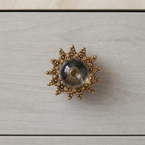 May include: A circular drawer knob featuring a central clear glass sphere surrounded by a decorative, sunburst-shaped brass-toned metal frame with intricate, embossed circular and floral patterns along its outer edge.