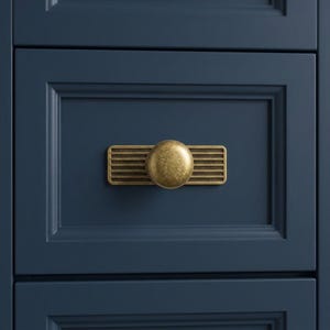 May include: A brass-toned cabinet knob featuring a circular center mounted on a rectangular backplate with horizontal grooved lines. The hardware is attached to a dark navy blue drawer front with a recessed panel and beveled edges.