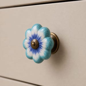 May include: A flower-shaped ceramic drawer knob featuring a white center with a blue starburst pattern, surrounded by teal petals. The knob is attached to a circular antique brass-toned metal base, providing a secure mounting point for furniture.