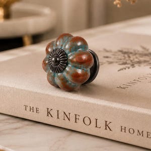 May include: A pumpkin-shaped cabinet knob featuring a mottled glaze of teal and rust-brown colors. The center of the knob has a dark, circular metal accent with a ridged, sunburst-like pattern, and a dark metal base connects it to the surface.