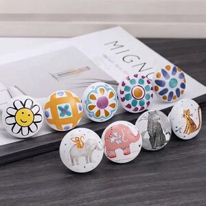 May include: A collection of eight round ceramic knobs featuring various designs including a yellow daisy with a smiley face, a yellow grid pattern, floral motifs, a gray elephant with a monkey, a pink elephant, a leopard, and a cheetah.