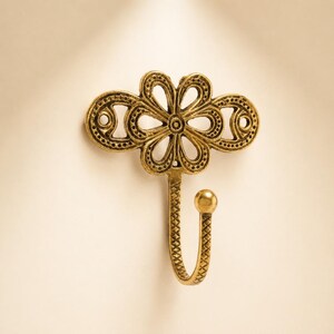 May include: A brass-colored metal wall hook featuring an ornate, openwork floral design at the top with two circular screw holes on the sides. The hook itself has a textured, cross-hatched pattern and ends in a smooth, rounded ball tip.