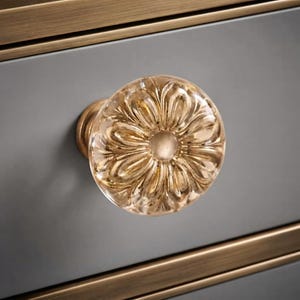 May include: A round, clear glass cabinet knob featuring a molded floral petal design. The knob is mounted on a circular brass-toned base and attached to a gray drawer front with horizontal brass-toned trim accents along the top and bottom edges.