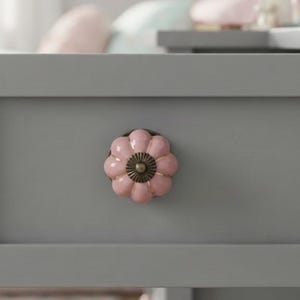 May include: A flower-shaped drawer knob with a glossy light pink finish and a central dark metallic circular accent. The knob is mounted on a flat, matte gray surface, featuring a scalloped edge design that mimics the appearance of a small bloom.