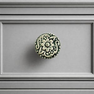 May include: A round cabinet knob featuring a raised, intricate floral and scrollwork pattern in a muted, weathered metallic finish. The design includes a central flower surrounded by small dots and curved lines, mounted on a flat gray drawer front.