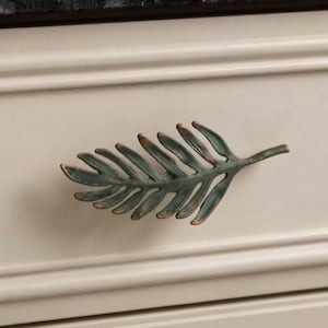 May include: A metal cabinet pull shaped like a fern leaf with a weathered, verdigris green and bronze finish. The handle features a detailed, textured vein pattern and is mounted horizontally on a smooth, light beige drawer front with molding.