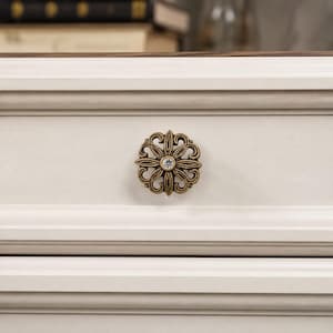 May include: A round, antique brass-toned metal drawer knob featuring an intricate, openwork floral or filigree pattern with a single, clear, faceted crystal stone set in the center, mounted on a smooth, off-white, horizontal wooden drawer front.