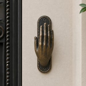 May include: A bronze-toned metal door handle shaped like an open human hand, mounted on a vertical oval backplate with a beaded edge. The hand is positioned with fingers pointing upward, secured to the wall by two visible screws at the top and bottom.