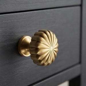 May include: A round, brushed brass cabinet knob with a fluted, sunburst-like radial pattern. The knob features a smooth, metallic finish and is mounted onto a dark gray, wood-grained drawer front, showcasing a clean, circular design profile.