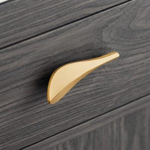 May include: A gold-toned, curved metal drawer pull with a smooth, polished finish attached to a dark gray, wood-grained cabinet drawer. The handle features an asymmetrical, tapered shape that extends horizontally across the textured surface.