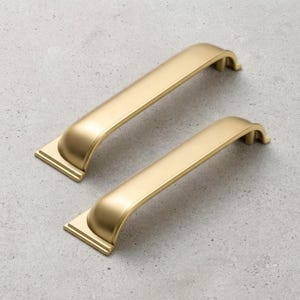 May include: Two gold-colored metal cabinet pulls with a smooth, arched design and rectangular base plates. Each handle features a curved grip section that transitions into a flat, stepped mounting bracket at both ends, finished in a matte gold tone.