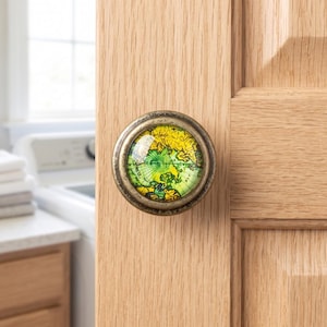 May include: A round cabinet knob featuring a vintage-style map design under a clear glass dome. The map displays yellow and green landmasses against a light background. The knob has a dark, weathered bronze-toned metal frame and circular base.