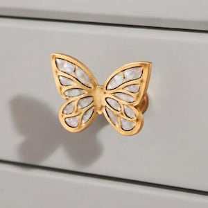 May include: A butterfly-shaped drawer knob featuring a gold-toned metal frame with intricate cutouts. The wings are inlaid with iridescent, pearlescent white segments that catch the light, creating a shimmering effect on the surface.