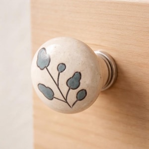 May include: A round, cream-colored ceramic cabinet knob featuring a hand-painted botanical design in muted blue-grey. The knob has a smooth, glossy finish and is attached to a silver-toned metal base, mounted onto a light-toned wooden surface.