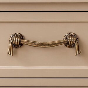 May include: A brass-toned metal drawer pull featuring a curved handle held by two sculpted hands at each end. The mounting plates are circular with decorative etched patterns, and the handle ends extend downward in a draped, fabric-like design.
