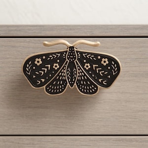 May include: A moth-shaped drawer knob featuring a matte black finish with gold-toned metallic outlines and detailing. The wings are decorated with embossed floral and geometric patterns, including small flowers, dots, and lines across the surface.