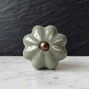 May include: A sage green ceramic cabinet knob shaped like a six-petaled flower. The knob features a smooth, glossy finish with a small, round, metallic bronze-colored center accent piece positioned in the middle of the floral design.