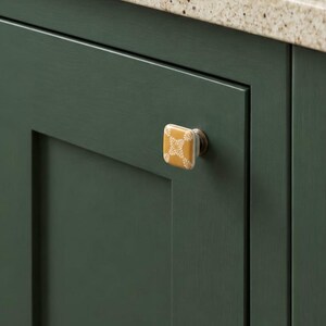 May include: A square cabinet knob with a mustard yellow finish and a white botanical leaf pattern, featuring a metallic base attached to a dark green cabinet door with a recessed panel design and a light speckled countertop edge.