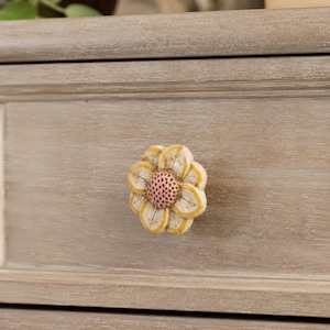 May include: A flower-shaped drawer knob featuring cream-colored petals with yellow outlines and a textured, light pink center. The knob is attached to a light brown, wood-grained drawer front with a horizontal panel design and a smooth finish.