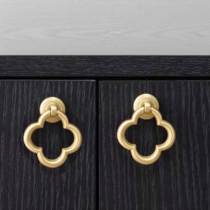 May include: Two gold-toned metal ring pulls with a quatrefoil shape mounted on dark black wood cabinet doors. Each pull features a circular base plate and a decorative clover-like ring that hangs vertically from a central hinge mechanism.
