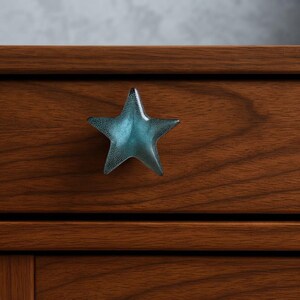 May include: A five-pointed star-shaped drawer knob with a textured surface in a gradient of deep teal to light blue, mounted on a horizontal wooden drawer front with a visible grain pattern and a dark brown finish.