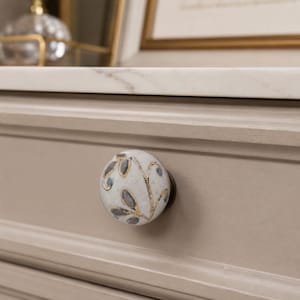 May include: A round ceramic drawer knob featuring a speckled off-white base with a hand-painted pattern of dark gray leaves outlined in metallic gold. The knob is mounted on a light beige wooden drawer front with a white marble countertop above it.