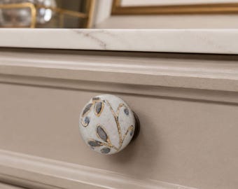 May include: A round ceramic drawer knob featuring a speckled off-white base with a hand-painted pattern of dark gray leaves outlined in metallic gold. The knob is mounted on a light beige wooden drawer front with a white marble countertop above it.