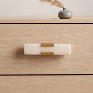 May include: A rectangular drawer pull featuring a smooth, off-white stone bar accented by two brushed gold metal brackets. The hardware is mounted horizontally on a light-toned wood drawer front, showcasing a clean, linear design with contrasting textures.